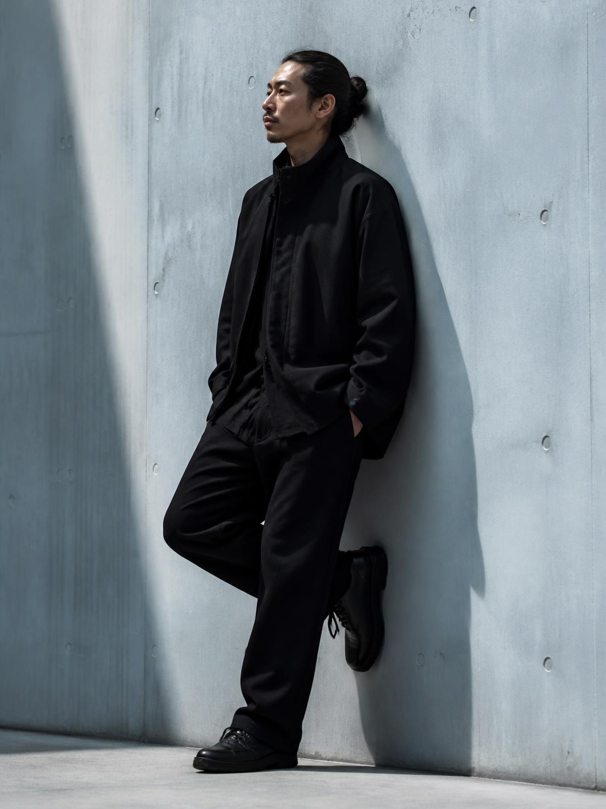 Full-length portrait of a man in all black against a pale blue-grey concrete wall